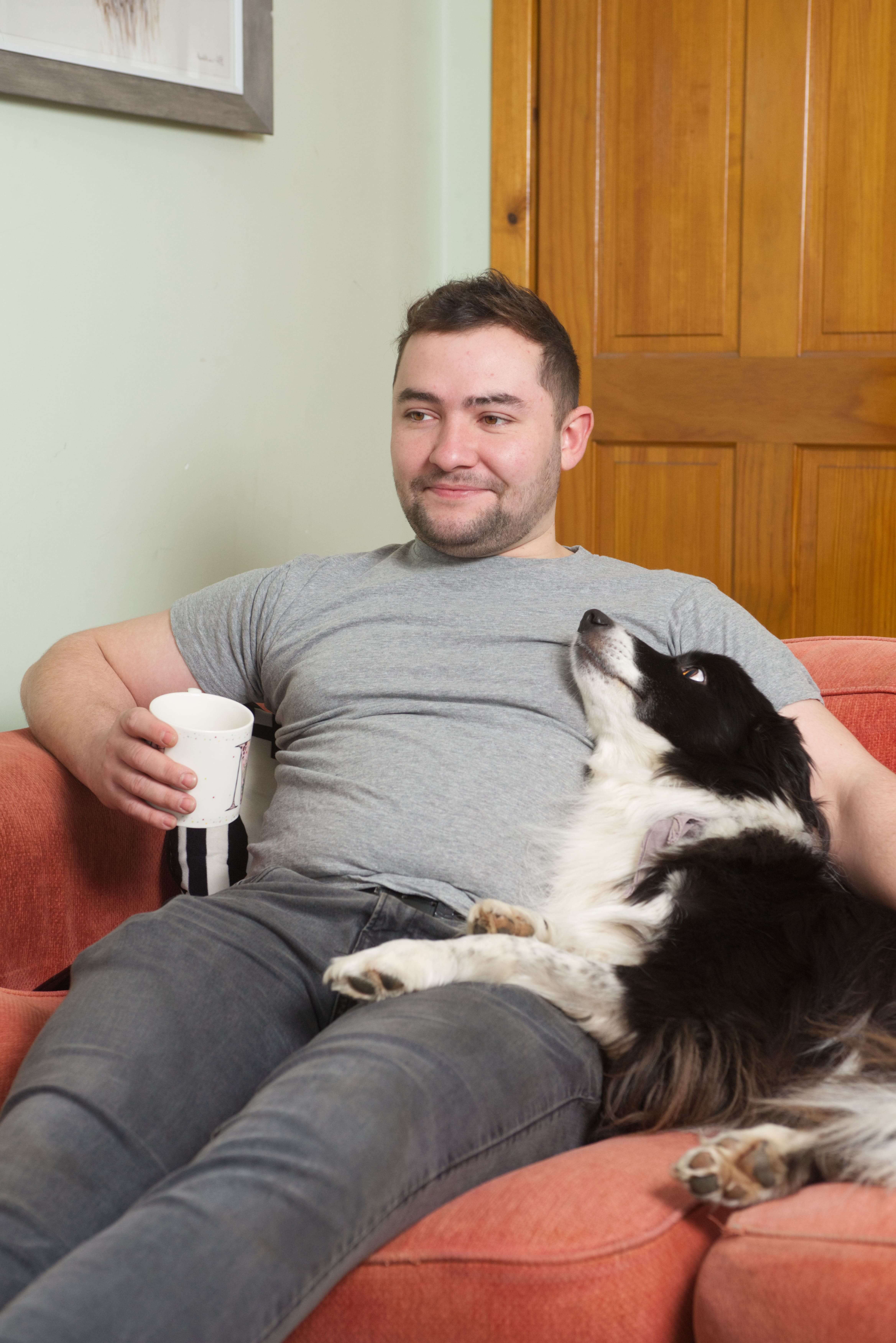 A man sits on his red sofa with a hot drink in a white mug. His black and white dog is sitting next to him and looking at him.