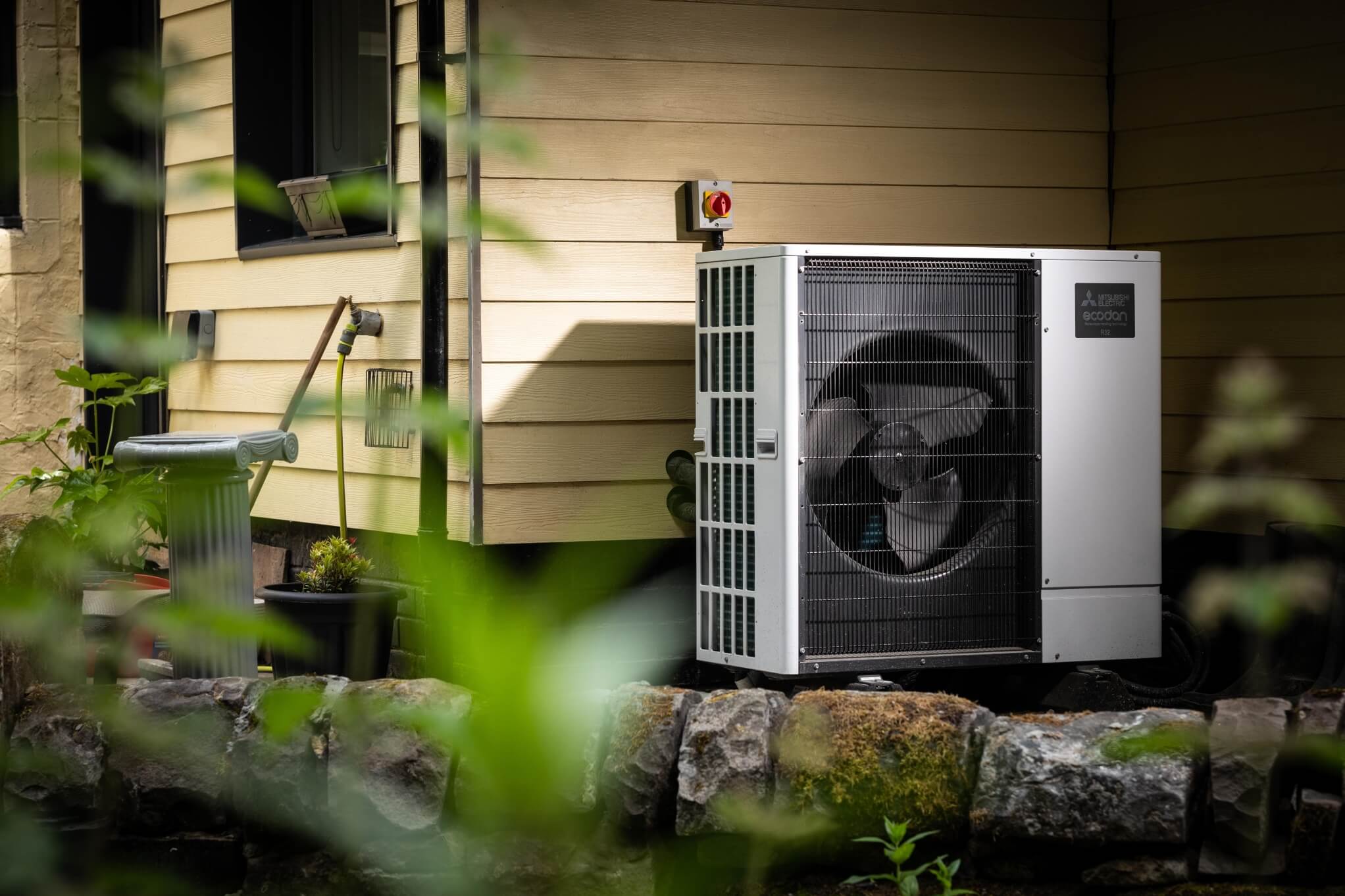 A heat pump against a beige wooden wall of a house house behind a low stone wall. Leaves from a tree cover the left hand side of the picture.
