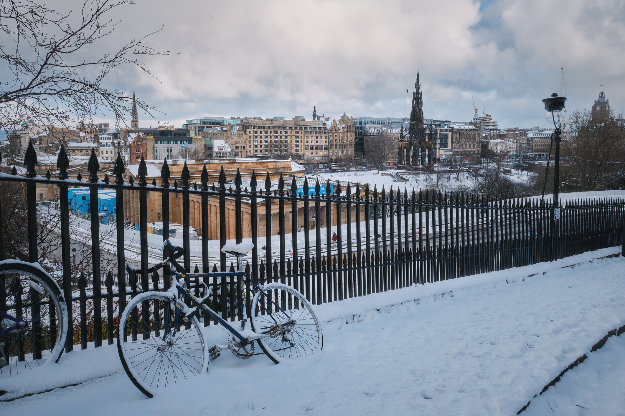 A bike is leaning against some railing in the snow. A wintery view of Edinburgh sits behind the railing.