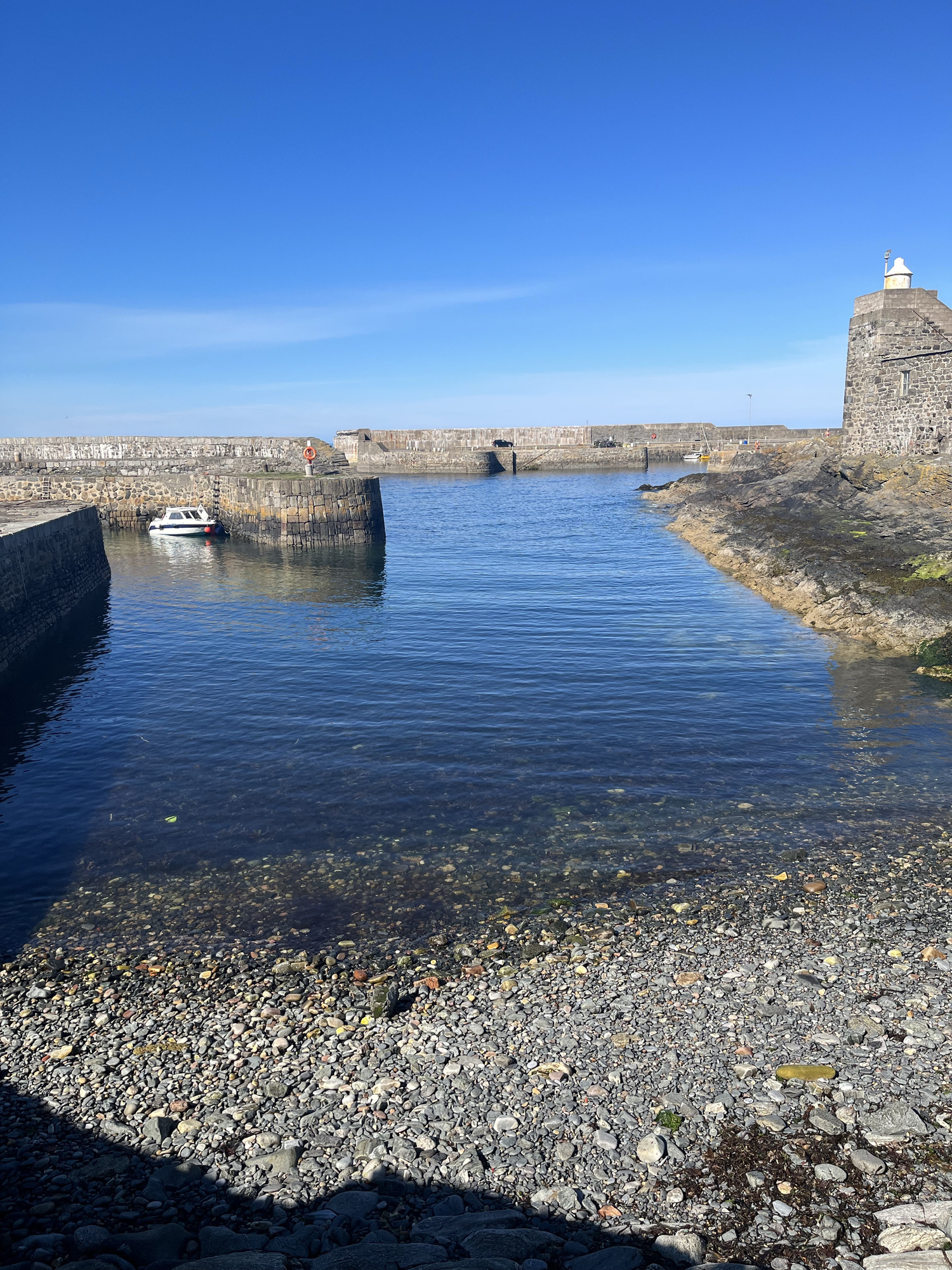 A view out from the shore of a harbour in Banff, Aberdeenshire. There is a clear blue sky and a there is a small medieval style stone building in the corner. 