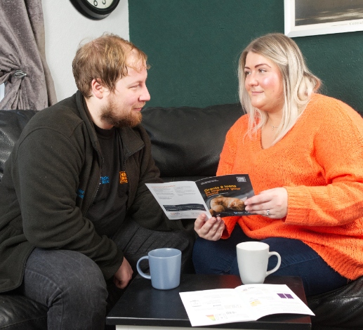 An advisor and householder sit on a couch looking at a Home Energy Scotland leaflet, there are cups of tea on the table in front of them