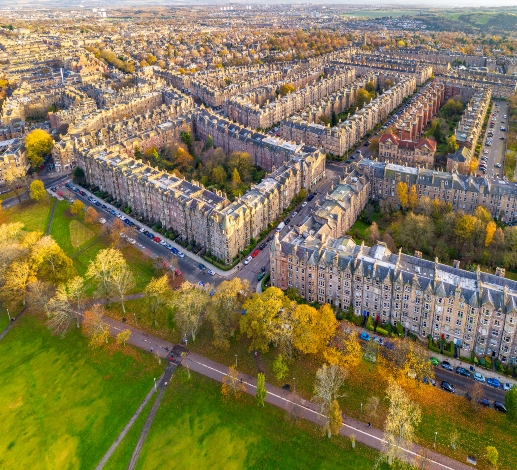 An aerial view of tenement blocks in Edinburgh on a sunny day. A line of autumnal trees can be seen in front of the tenements, as well as the green grass of a park with a footpath running along its center. 