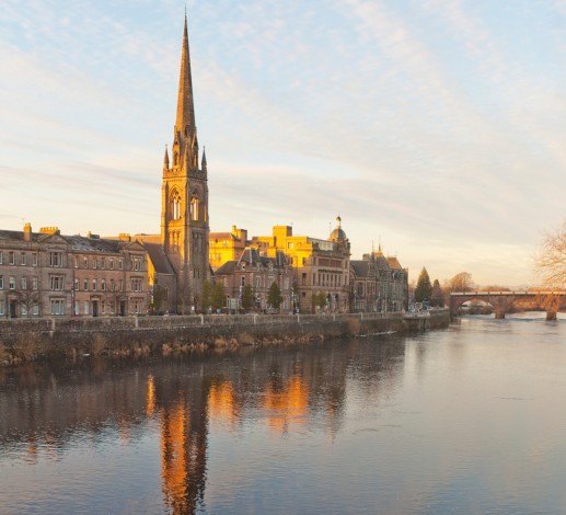 Buildings along the River Tay in Perth.  A bridge can be seen going across the river. Sunlight is hitting the rooftops in the center of the image.  