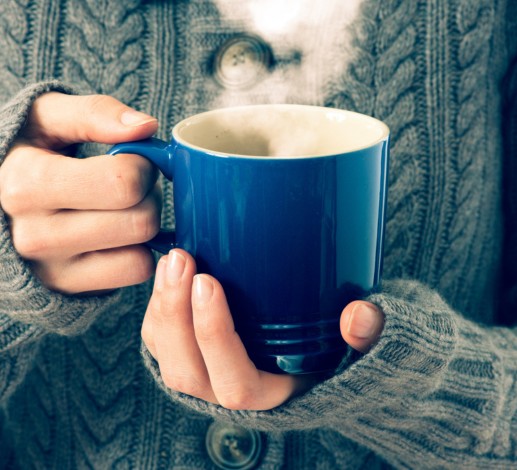 A person wearing a grey knitted cardigan is holding a blue ceramic mug in both of their hands, steam can be seen coming from the mug.  