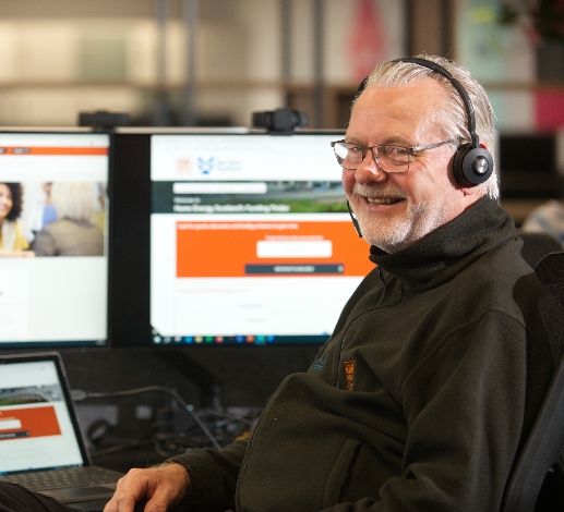 Harry, a Home Energy Scotland advisor, is at a desk and computer wearing a telephone headset, he is smiling