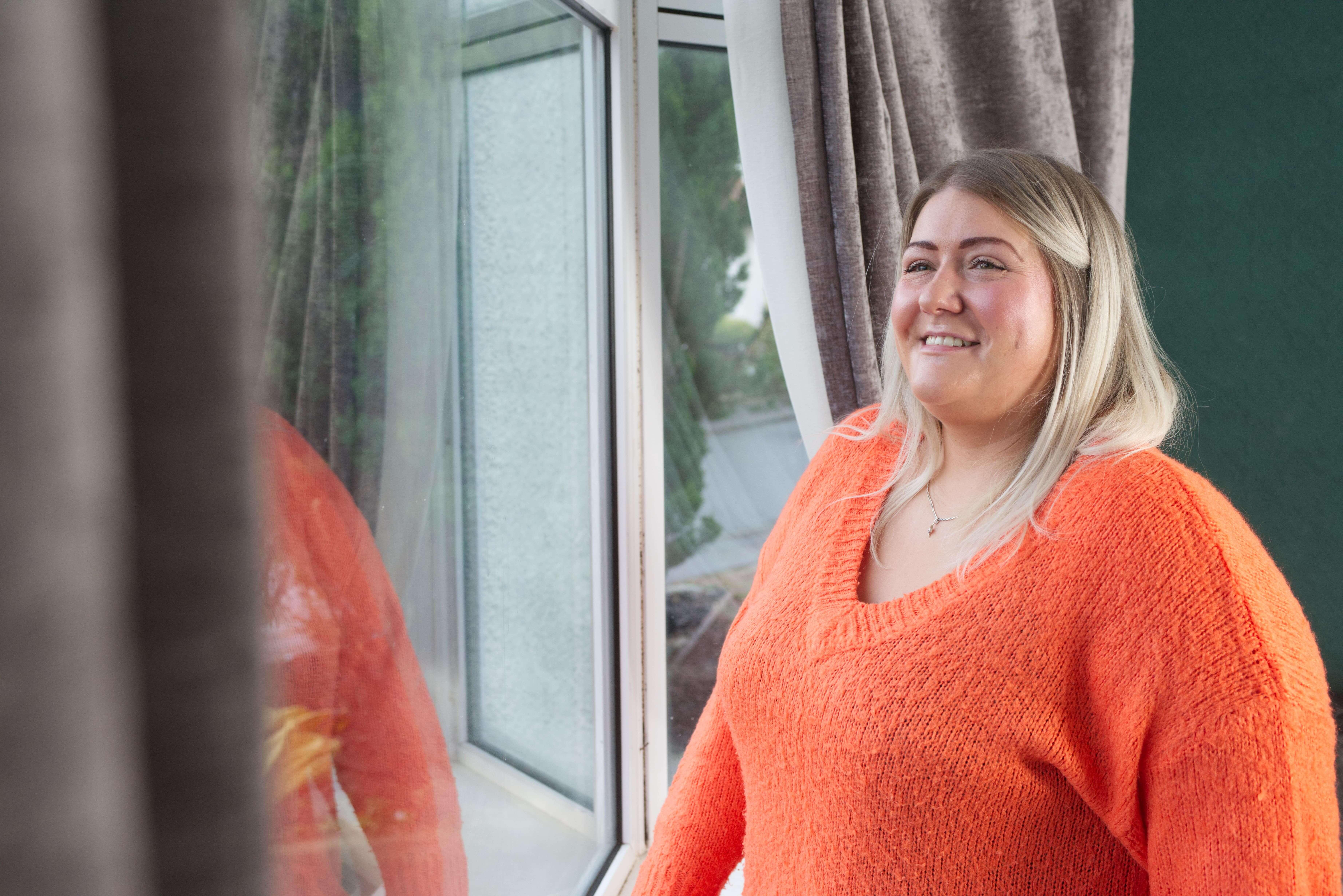 Woman wearing bright orange jumper smiles while looking out of her double glazed window